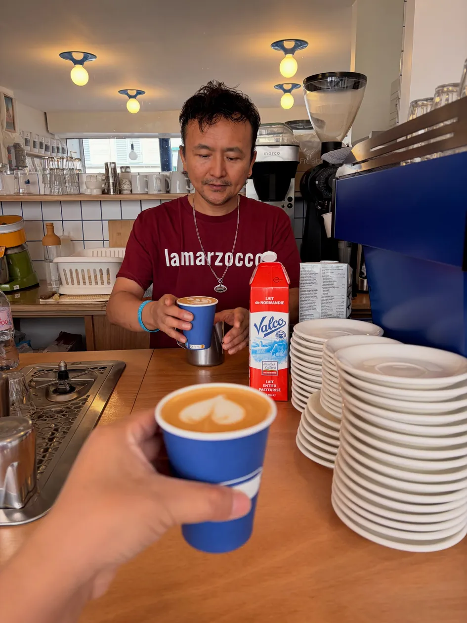 Monlam au comptoir, à côté de la machine bleue, tend un café dans un gobelet bleu
